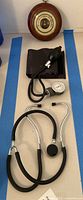 Photo showing sphygmomanometer, stethoscope and round barometer on blue taped surface.