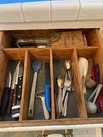 View of kitchen drawer with wooden organizer containing four wood handled knives, assorted metal and plastic cooking utensils including spatula and can opener, silver spoons and forks, and white plastic spoons.