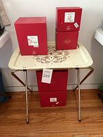 Red Lenox boxes labeled with holiday-themed decorative sets atop a cream colored vintage TV tray with a dark brown floral design, standing on a wooden floor.