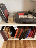 Shelf containing Panasonic VHS player, VHS tapes, some books, and cardboard boxed VHS tapes.