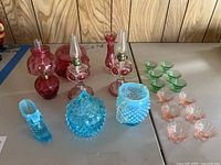 Photo showing full assortment of vintage glass items including pink glass oil lamps, blue hobnail glass containers, shoe figurine, and various small glass bowls