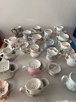 Front overview of 21 vintage porcelain men's shaving mugs arranged on table showing variety of designs and pastel colors with floral decorations and gold trims