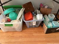 Full shot showing three bins and one box filled with assorted vintage Tupperware and miscellaneous kitchen items on wooden floor.