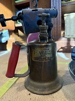 Side view of one antique camping burner with red handle and caution label, showing rust and patina