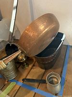 Photo showing hammered copper pan from side angle alongside metal sifter, small decorative metal tub, and brass candle holder on wooden surface.