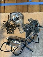 Photo of one antique chrome handheld electric hair dryer with vent slots, and two vintage electric clothing irons with cords on beige carpet.