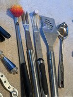 Various metal kitchen utensils including spatulas with black handles, large metal spoon, and fork for cooking or BBQ use