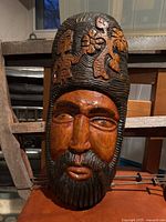 Front view of the hand-carved wooden mask showing detailed carvings of a man's face with beard and floral decorations on the headpiece.