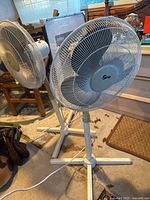 Two standing fans in a room, one white and blue Ozeri fan and one Beige Ocean Breeze fan, both with power cords visible on the floor