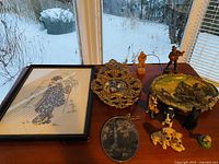 View of framed stitchwork art, filigree wall art, plaster cityscape plaque, dog figurines, and carved wood figurines on wooden table near window with snowy outdoors