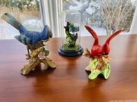 Front and side view of blue jay and cardinal ceramic bird figurines with enclosed bluebird dome beside them on wooden table near window