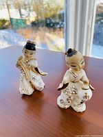 Front view of two small ceramic dancing children figurines, one holding a fan and the other holding a lantern, placed on a wooden surface with outdoor background visible.