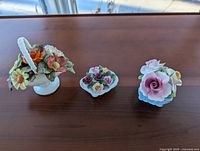 Three porcelain floral china pieces placed on wooden surface: blue basket with assorted roses, white seashell-shaped piece with roses, white handled basket with assorted flowers.