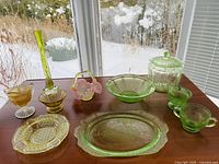 Photo of all colored glass Depression glass dishes arranged on a wooden table near window with snow outside, highlighting green and yellow pieces including tall vase, jar with lid, bowls, cups, and platters.