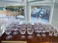 Full set of crystal stemware arranged on a table by the window with snowy outdoor view, showing 7 large water glasses, 8 champagne glasses, 8 wine glasses, and 8 sherry/cognac glasses all made of clear faceted crystal.