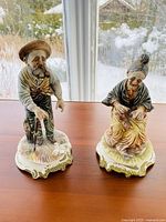 Front view of two elderly person figurines, man standing with cane and woman sitting knitting, on a wooden table by a window with snowy outdoors.