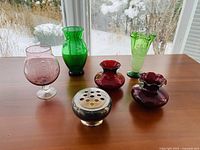 Six multi-coloured glass vases and one flower frog arranged on wooden table with snow outside window