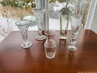 View of five clear glass flower vases with twisted and textured bottoms, standing on a wooden surface by a window with snow outside.
