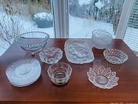Photo showing full set on wooden table including large footed bowl, smaller bowls, rectangular serving tray, plates, and small dishes in clear glass with floral and geometric patterns.