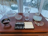 Full view of all glassware items arranged on wooden table including casserole dish with lid, water pitcher, dessert plates, small bowls, candy dish, honey pot, and boxed dessert forks.