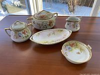 Photo showing the full set of five pieces in the hand painted tea service, placed on a wooden surface with outdoor background.