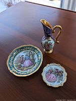 Photo showing all three items: one miniature vase with curved handle and two plates, on a wooden surface.