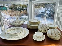 Full overview of all china pieces arranged on table including plates, cups, and serving dishes with window and snowy view in background.
