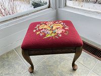Front angle showing the red needle point floral upholstery and carved wood legs of the foot stool