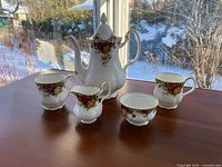 Front view of the entire 5-piece tea service set on a wooden surface near window light, showing the teapot, two mugs, creamer, and sugar bowl, all decorated with the floral Old Country Roses pattern and gold trim.