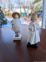 Two Royal Doulton porcelain figurines, girl in nightgown and boy with straw hat and dog, shown on wooden table with window background.