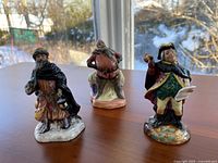 Front view of the three Royal Doulton figurines Good King Wenceslas, Town Crier, and Falstaff on a table with a snow-covered window background.