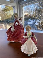 Two Royal Doulton figurines placed on a wooden table by a window with natural light showing details of the dresses and faces.