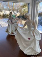 Photo showing both figurines on display with winter landscape background; one standing 'Country Rose' with floral dress and one seated 'Tracy' in white flowing dress