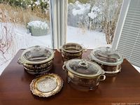 Four silver serving dishes each with a clear glass casserole dish lid and two silver butter trays displayed on a wooden table, in natural light showing some tarnish on silver needing polishing.