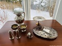 View of all silver plated items on wooden table by window including water jug, cream and sugar containers, salt and pepper shakers, hot plate, tier tray, and napkin ring.