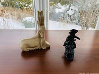 Photo showing both sculptures on a wooden surface near a window with snow visible outside: the stone goat carving on the left and the metal kids sledding sculpture on the right.
