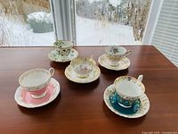 Overview of 5 teacups and matching saucers arranged in a tight group on wooden table with snow visible outside, showing patterns and gold trim on each set.