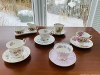 Five floral teacups with matching saucers displayed alongside matching creamer and sugar bowl on a wooden surface near a window with snowy outdoor view.