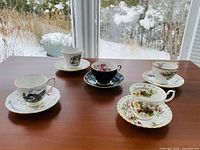Teacups and saucers displayed on wooden table by window with snow outside. Various designs visible including black exterior with floral interior and house and landscape scenes.