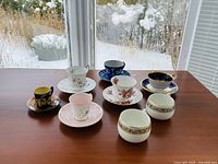 Set of 6 teacups and saucers plus 2 small cups arranged on a wooden table near a snowy window, showing varied floral and decorative patterns.