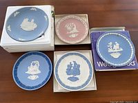Overview of five Wedgwood commemorative plates showing variety of blue and pink jasperware with white relief designs. Plates are presented on boxes and foam pads.