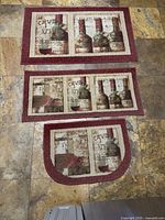 Three kitchen rugs arranged on floor: large rectangular, medium rectangular, and small U-shaped with wine bottle and glass designs in burgundy and earth tones.