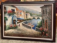 Full view of the framed oil painting showing an Italy canal scene with boats, stone buildings, flowers and mountains in the background.