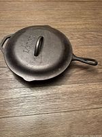 Cast iron skillet with lid placed on wood surface, showing 'Lodge' embossed on lid handle and overall shape.