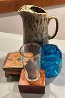 Photo showing all four items of the lot on tabletop: Nudie drip pottery pitcher, Florentine Italy lidded box, cedar sample box, blue glass jar with lid.