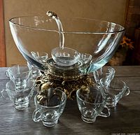 Front view of punch bowl set with bowl on ornate gold stand surrounded by 9 clear glass cups and the ladle inside the bowl