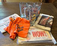 Photo of six tall Arby's Denver Broncos beer glasses with football bases, an autographed orange Broncos scarf from 1988, and vintage Denver Broncos newspapers on a wooden table.