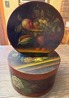 Large round wooden box with a hand-painted fruit basket still life on the lid, resting on smaller box painted with floral design