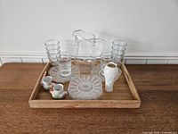 Full view of the breakfast set including juice jug, 12 glasses stacked in two piles, 8 dishes laid flat, 2 decorative egg cups, Lenox bud vase, and wooden tray.