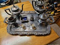 Full view of the ten silverware items arranged on a large ornate rectangular silver tray on a wooden table.